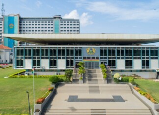 Modern government building with a wide staircase, flanked by greenery, and a central emblem above the entrance.