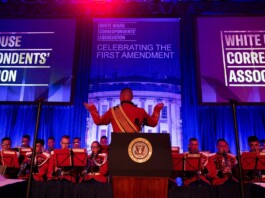 Conductor raises arms to lead a red-uniform band performing on a stage with White House Correspondents' Association banners behind.