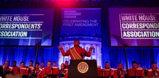 Conductor raises arms to lead a red-uniform band performing on a stage with White House Correspondents' Association banners behind.