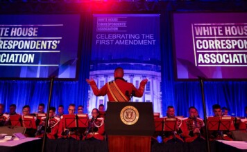 Conductor raises arms to lead a red-uniform band performing on a stage with White House Correspondents' Association banners behind.