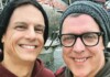 Two men outdoors taking a selfie, smiling, wearing knit beanies near a fountain behind them.
