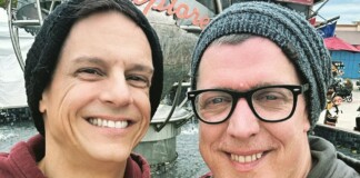 Two men outdoors taking a selfie, smiling, wearing knit beanies near a fountain behind them.