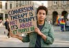 Person in a denim jacket holds a cardboard protest sign that reads, 'Conversion therapy is not therapy. It's abuse. Let people be' in a city square.