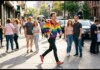 Young man in a rainbow jacket strides across a busy city street as onlookers watch from the sidewalk.