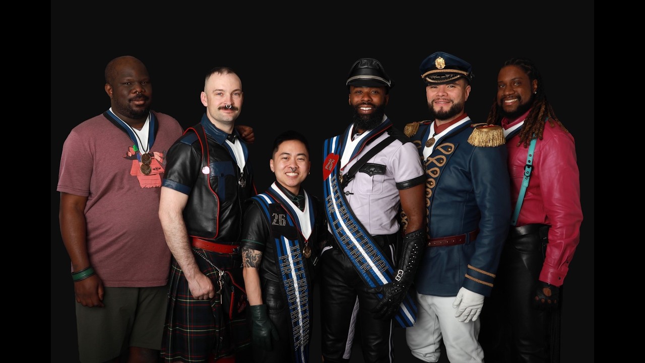 Honey Davenport Makes History With International Mr. Leather Win Group portrait of six men in colorful leather outfits and ceremonial sashes posing together against a black backdrop, smiling at the camera.