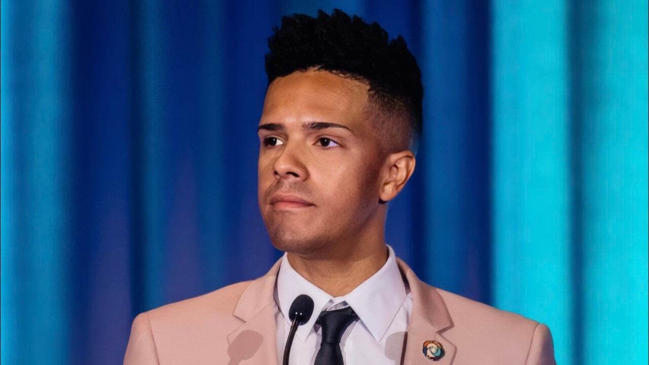 Young man in a light pink blazer speaking at a microphone on a stage with blue curtains behind him.
