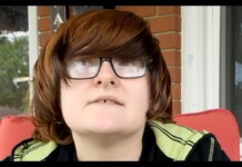 8th Grader’s Graduation Speech Accusing School of ‘Homophobia’ Goes Viral Online Person with shoulder-length brown hair and large glasses sits on a red outdoor chair on a brick porch, nose ring visible.