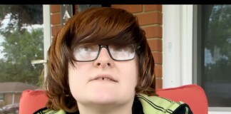 Person with shoulder-length brown hair and large glasses sits on a red outdoor chair on a brick porch, nose ring visible.