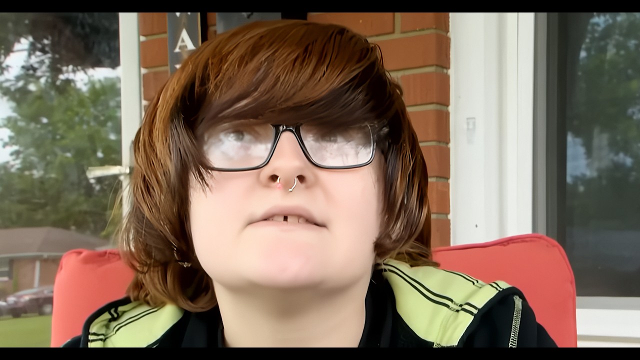 Person with shoulder-length brown hair and large glasses sits on a red outdoor chair on a brick porch, nose ring visible.