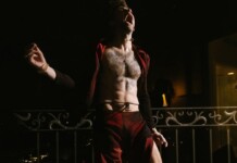 Beartooth Singer Caleb Shomo Comes Out as Gay Following 14-Year Marriage Shirtless man shouting on a balcony at night, wearing red pants and an open red jacket.