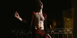 Shirtless man shouting on a balcony at night, wearing red pants and an open red jacket.