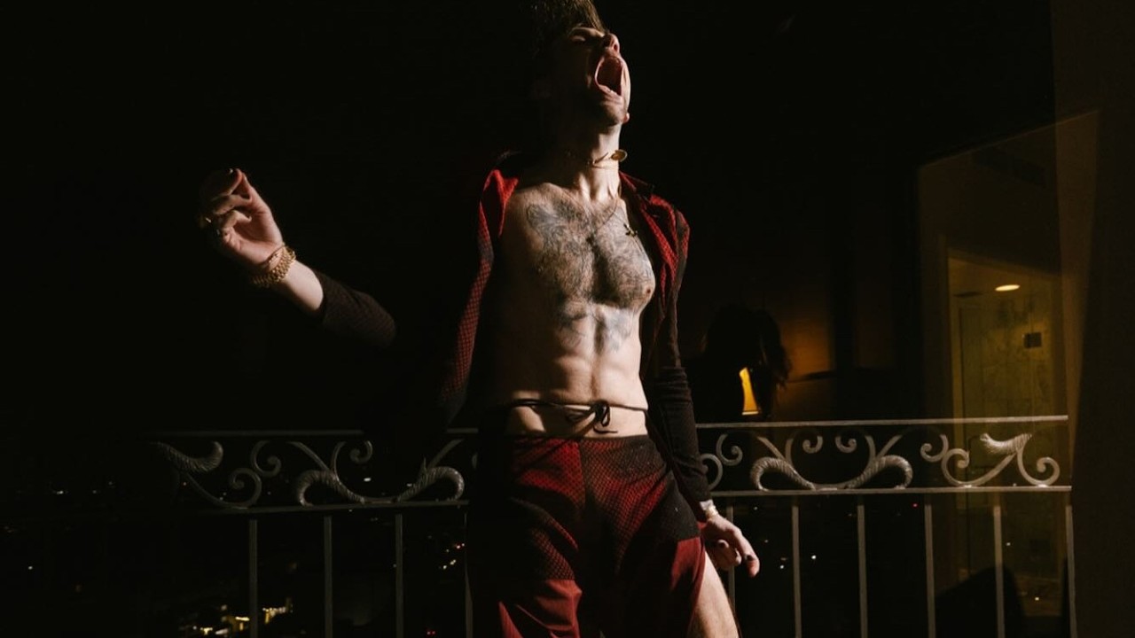 Shirtless man shouting on a balcony at night, wearing red pants and an open red jacket.