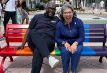 Two smiling people sit on a rainbow-colored park bench in a sunny urban plaza.