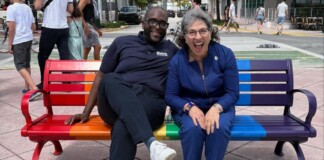 Two smiling people sit on a rainbow-colored park bench in a sunny urban plaza.