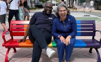 Two smiling people sit on a rainbow-colored park bench in a sunny urban plaza.