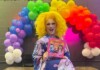 Michigan City Council Votes to Remove Drag Story Hour From Pride Festival Drag queen with bright yellow wig and colorful outfit sits in front of a rainbow balloon arch, holding a pink book smiling at the camera.