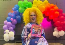 Michigan City Council Votes to Remove Drag Story Hour From Pride Festival Drag queen with bright yellow wig and colorful outfit sits in front of a rainbow balloon arch, holding a pink book smiling at the camera.
