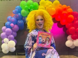Michigan City Council Votes to Remove Drag Story Hour From Pride Festival Drag queen with bright yellow wig and colorful outfit sits in front of a rainbow balloon arch, holding a pink book smiling at the camera.