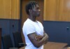 Six Flags Permanently Bans Teen Accused of Publicly Exposing Trans Woman at Cedar Point Young man with dreadlocks and a white t-shirt stands with arms crossed in a courtroom, looking to the right with a serious expression.