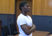 Six Flags Permanently Bans Teen Accused of Publicly Exposing Trans Woman at Cedar Point Young man with dreadlocks and a white t-shirt stands with arms crossed in a courtroom, looking to the right with a serious expression.