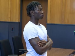Six Flags Permanently Bans Teen Accused of Publicly Exposing Trans Woman at Cedar Point Young man with dreadlocks and a white t-shirt stands with arms crossed in a courtroom, looking to the right with a serious expression.