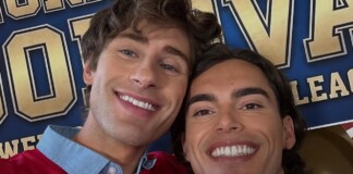 Two men smiling for a selfie in front of a blue and gold banner at an event.