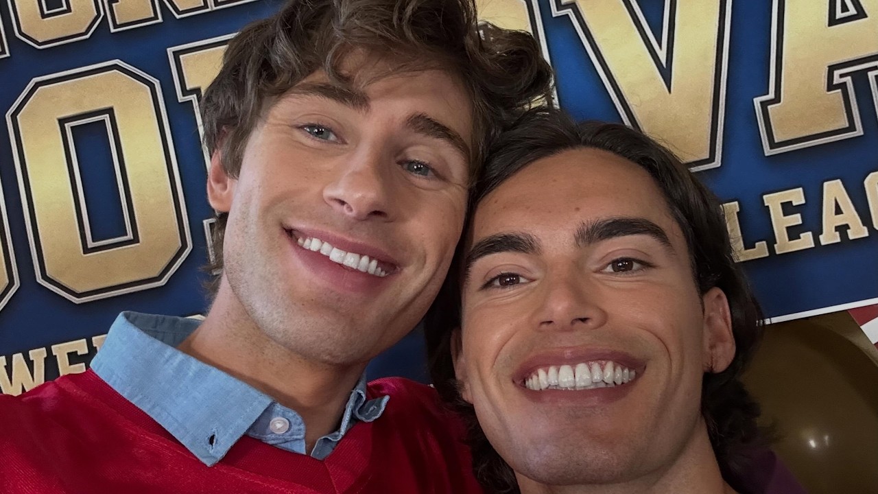 Two men smiling for a selfie in front of a blue and gold banner at an event.