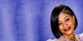 Portrait of a smiling woman with a black bob haircut, wearing layered necklaces and pearl earrings, against a purple backdrop.