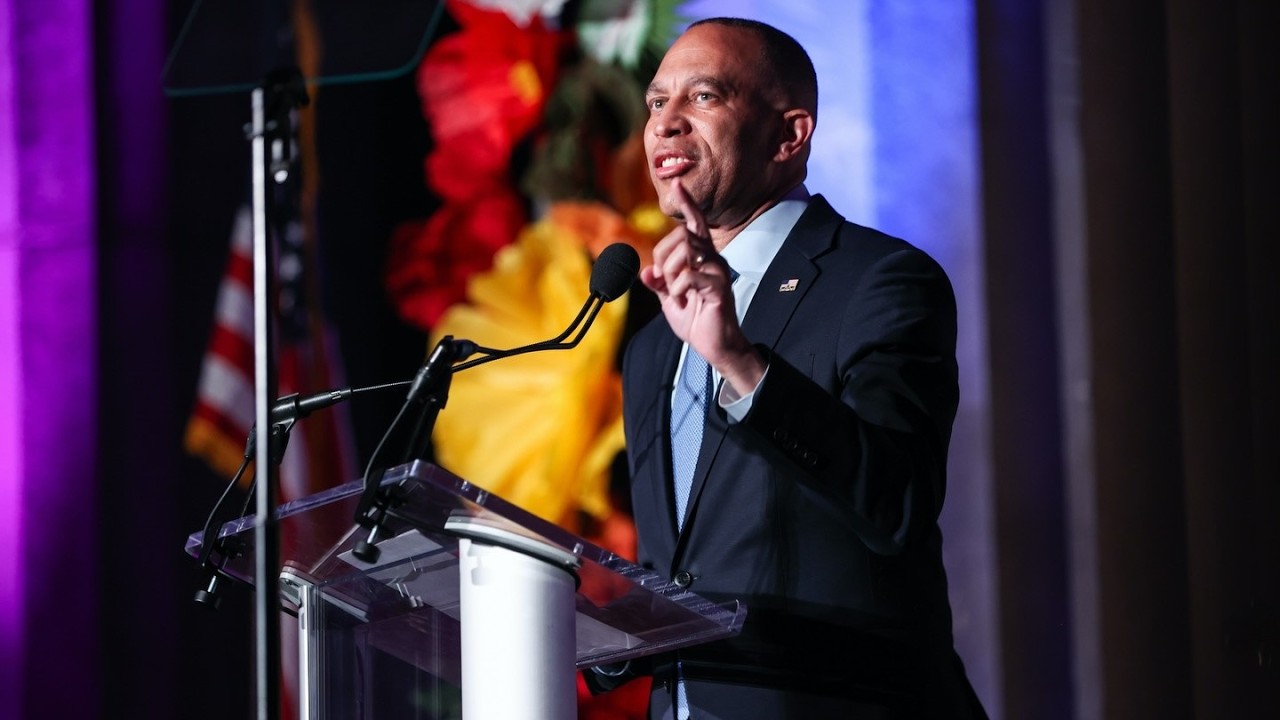 House Democratic Leader Hakeem Jeffries Promises To Stand With LGBTQ+ ‘Always and Forever.’ Man in a dark suit speaks at a clear podium on a stage with a microphone, gesturing with his hand against a colorful backdrop.