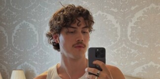Young man in a white tank top taking a mirror selfie with earphones in a wallpapered bedroom