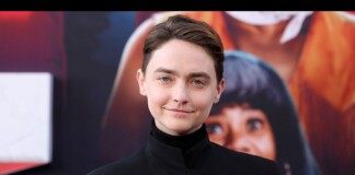 Person with short brown hair in a black high-collar outfit posing in front of a colorful poster backdrop.
