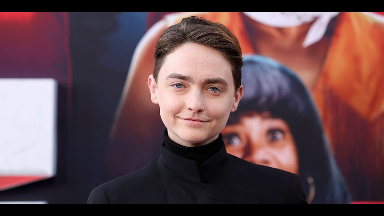 Person with short brown hair in a black high-collar outfit posing in front of a colorful poster backdrop.