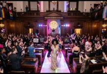 Drag queen in a glittering gown walks a runway inside a grand city hall chamber, surrounded by cheering attendees and Pride banners nearby.