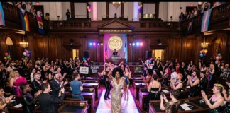 Drag queen in a glittering gown walks a runway inside a grand city hall chamber, surrounded by cheering attendees and Pride banners nearby.