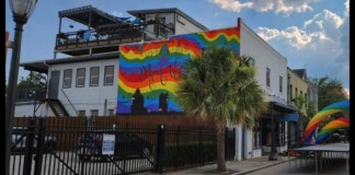 Rainbow mural on a white building reading 'Love Wins' with a palm tree in front and parked cars behind a black fence.