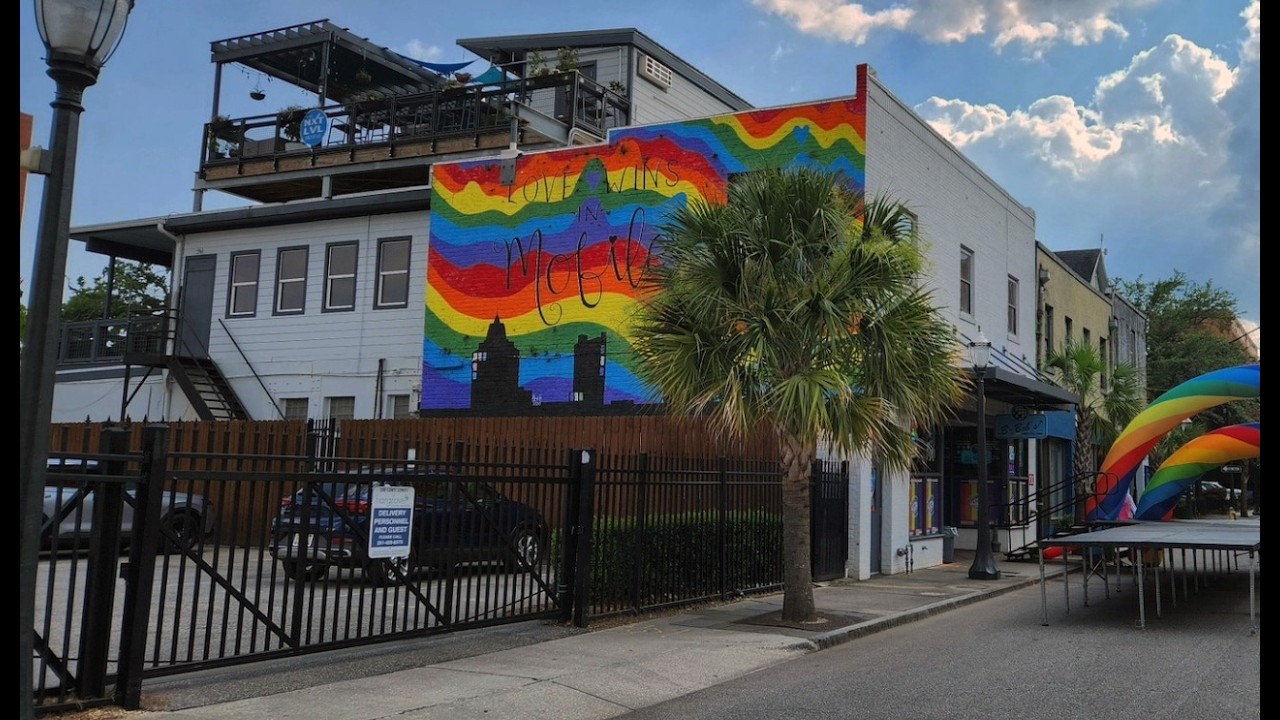 Rainbow mural on a white building reading 'Love Wins' with a palm tree in front and parked cars behind a black fence.