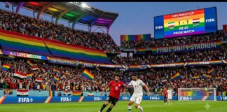 Soccer players contest the ball on a green pitch as a rainbow-filled crowd cheers in Pride Unity Stadium during the FIFA World Cup 1-1 Iran vs Egypt.