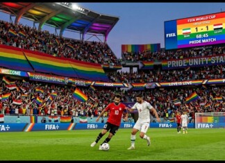 Soccer players contest the ball on a green pitch as a rainbow-filled crowd cheers in Pride Unity Stadium during the FIFA World Cup 1-1 Iran vs Egypt.