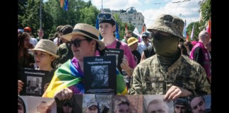 Crowd of protesters holding photos of missing people; a person wrapped in a Ukrainian flag stands beside a masked soldier in camouflage in an urban square.