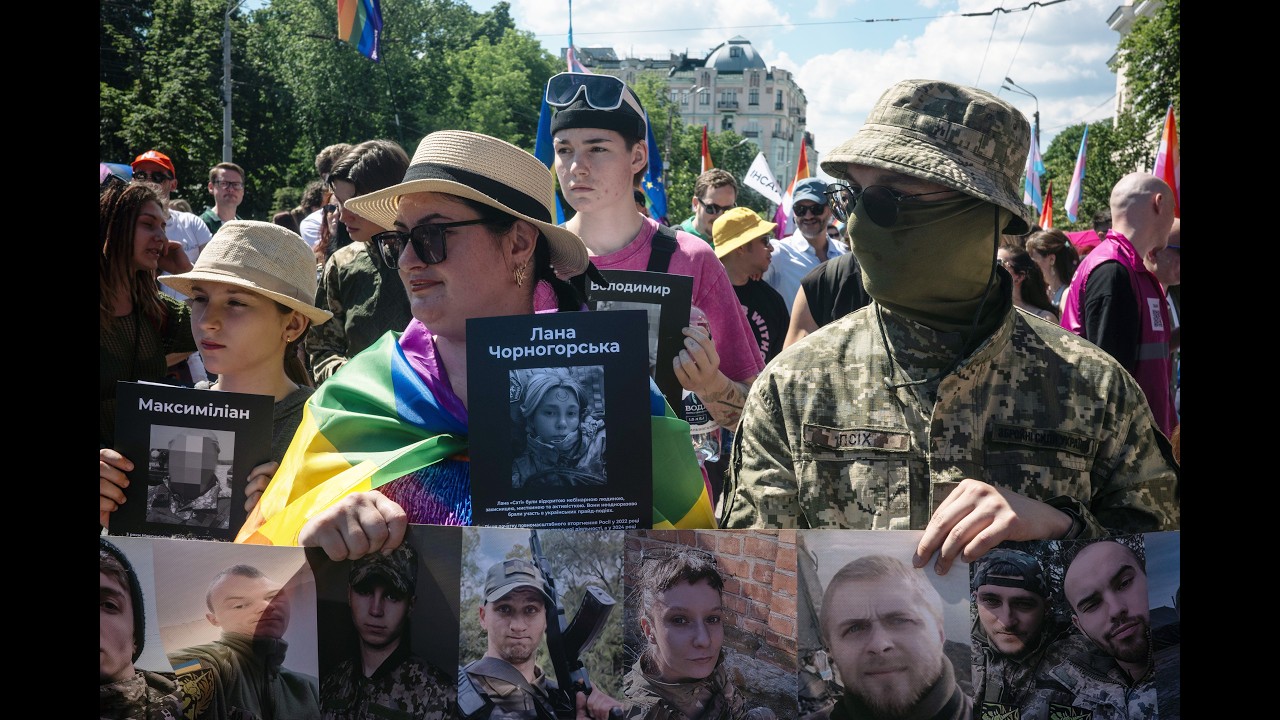 Crowd of protesters holding photos of missing people; a person wrapped in a Ukrainian flag stands beside a masked soldier in camouflage in an urban square.