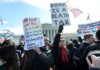 Crowd of protesters holding signs in front of a government building, including one that reads 'Doma is a Death Tax' with American flags nearby.