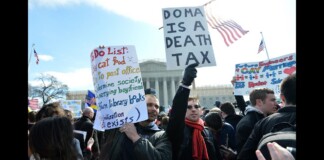 Crowd of protesters holding signs in front of a government building, including one that reads 'Doma is a Death Tax' with American flags nearby.