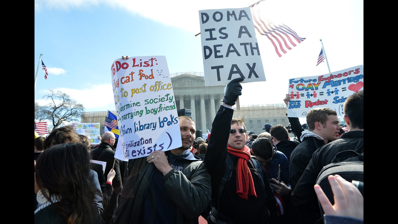 Crowd of protesters holding signs in front of a government building, including one that reads 'Doma is a Death Tax' with American flags nearby.