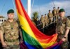 Military personnel in camouflage hold a large rainbow Pride flag outdoors.