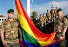 Military personnel in camouflage hold a large rainbow Pride flag outdoors.