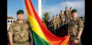 Military personnel in camouflage hold a large rainbow Pride flag outdoors.