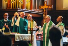 Priests in green liturgical vestments celebrate Mass at the altar; one priest elevates the Eucharist as others assist, with a crucifix and organ pipes in the background.