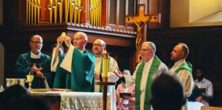 Priests in green liturgical vestments celebrate Mass at the altar; one priest elevates the Eucharist as others assist, with a crucifix and organ pipes in the background.