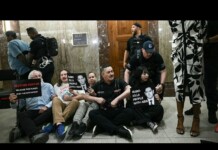 Protesters sit on the floor in a marble lobby, holding signs about funding and AIDS-related accusations as police stand nearby.