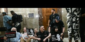 Protesters sit on the floor in a marble lobby, holding signs about funding and AIDS-related accusations as police stand nearby.